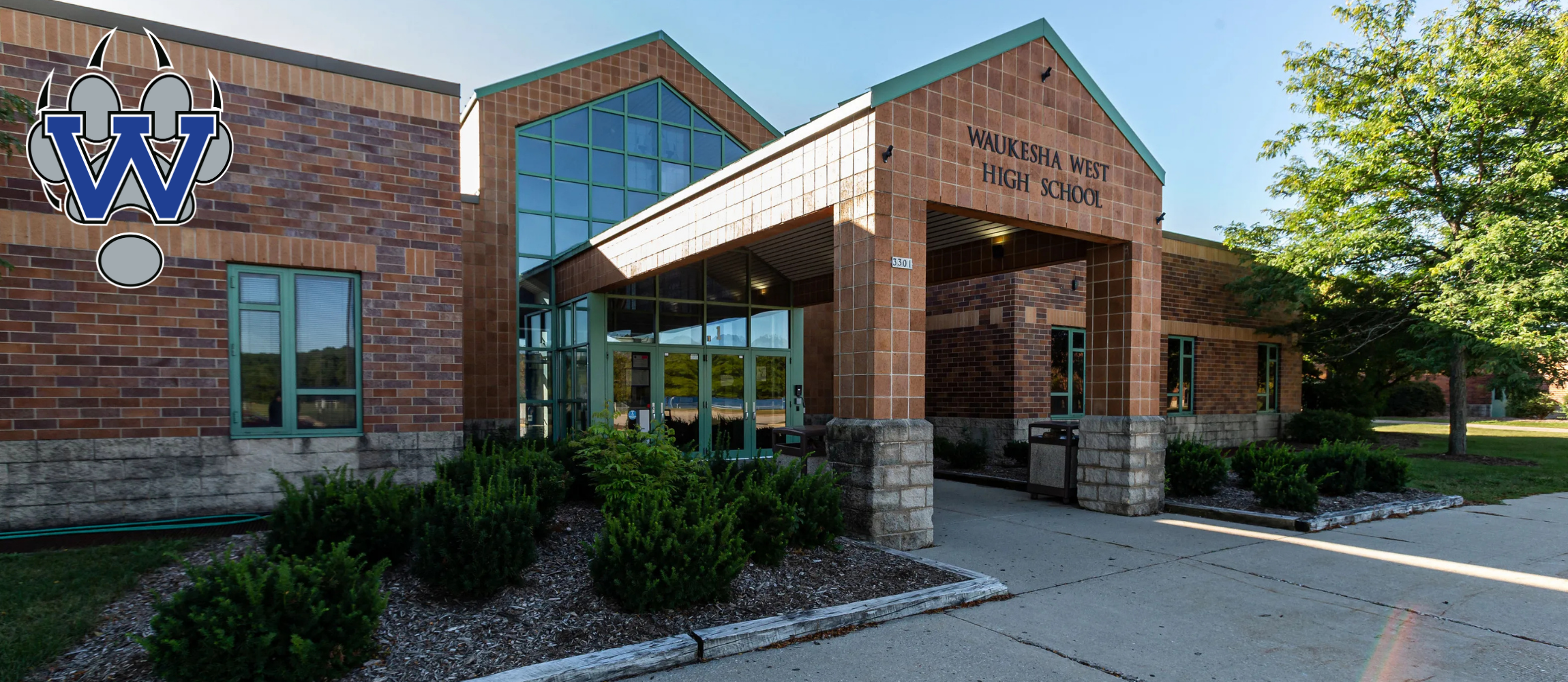 A building with brick walls and glass windows features an entrance with a canopy and logo. Surrounding it are plants and a driveway.