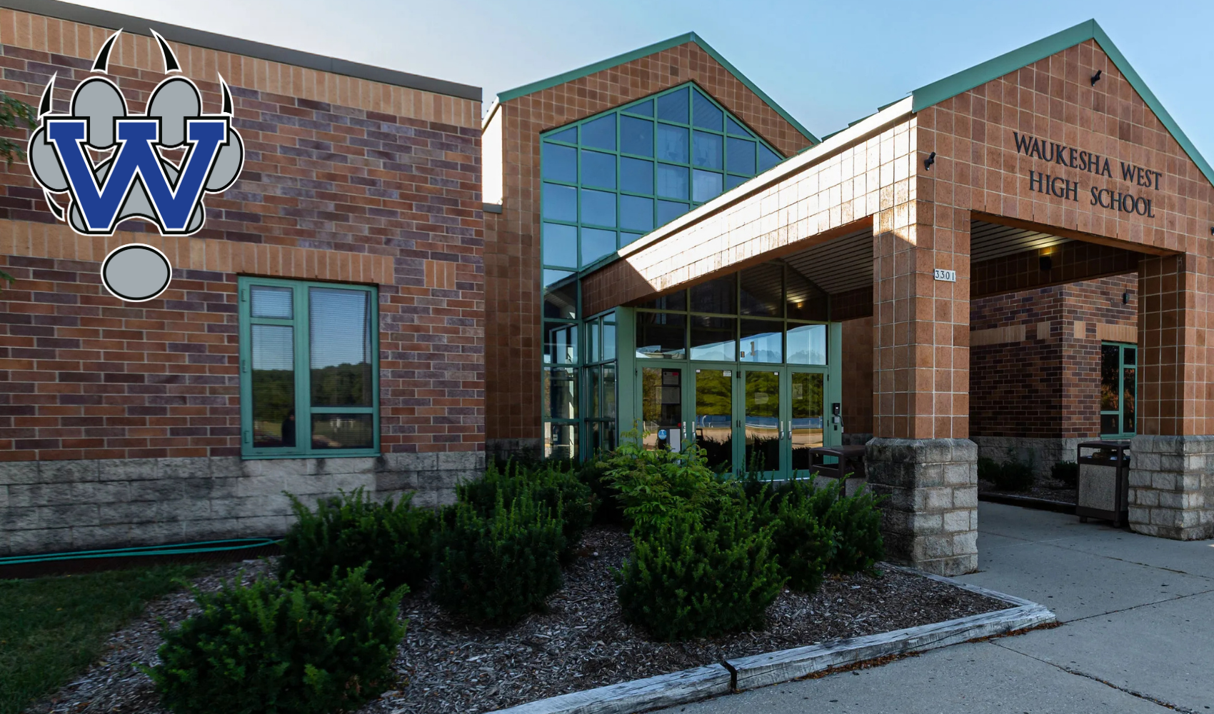 A building with brick walls and glass windows features an entrance with a canopy and logo. Surrounding it are plants and a driveway.
