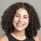 A woman with curly hair and braces smiles in a studio, wearing a black sleeveless top.