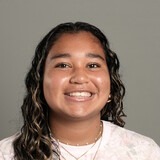 A woman with curly hair, wearing a white shirt with floral print, smiles for a portrait against a gray background.
