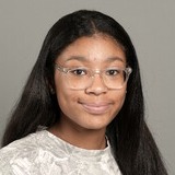 A woman with long, dark hair and glasses smiles at the camera. She wears a white, patterned shirt against a plain gray background.