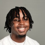 A smiling person with a beard and braided hair wearing a white t-shirt and a necklace.