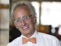 An older man with gray hair, wearing glasses and a white shirt with an orange bow tie, smiling.