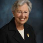 Woman in a black blazer and glasses smiles for a portrait against a blue background.