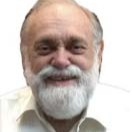 An older man with white hair, a beard, and mustache wearing a white shirt, smiling.
