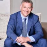 A man in a blue suit and tie sits on a light-colored couch, smiling at the camera.