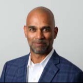 A man with a beard and a smile is standing in front of a plain background. He wears a white shirt and blue blazer.