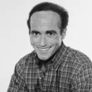 Black-and-white portrait of a smiling man with short, curly hair, wearing a plaid shirt. He is sitting down.