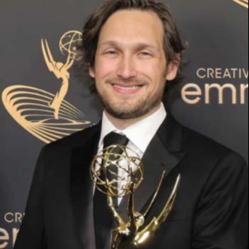 Man with a beard holding a gold Emmy statue, wearing a black suit and tie.