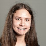 A woman with long brown hair and a smile wearing braces poses against a plain, gray background.