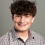 A smiling person with curly hair and a checkered shirt, wearing a gray background.