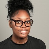 A woman with black hair in a bun, wearing glasses and a black shirt, smiles at the camera.