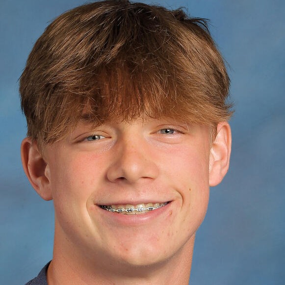 A boy with short brown hair and braces smiles, wearing a blue shirt and a silver necklace.