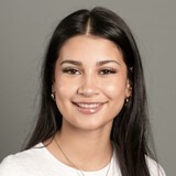 A person with long dark hair, wearing a white shirt and a necklace, smiles at the camera.