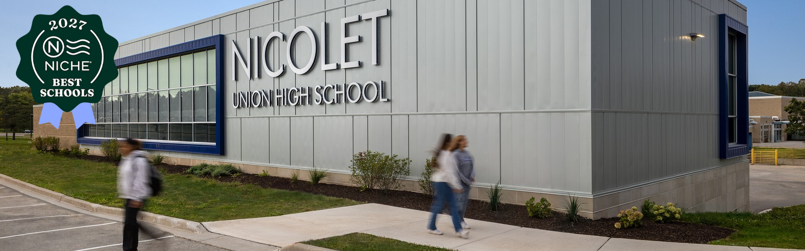 A modern building with "Nicolet High School" on the wall. Two individuals walk on the sidewalk.