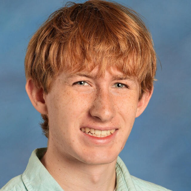 A young person with short, wavy, auburn hair smiling at the camera. They wear a light blue collared shirt against a blue background.