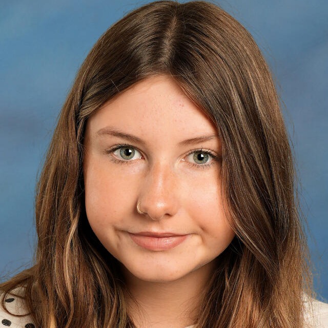 A young person with long, wavy brown hair smiles at the camera, wearing a white shirt with black polka dots.