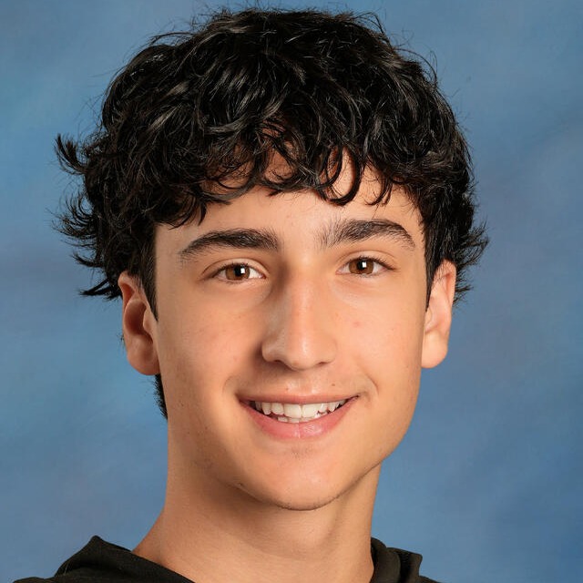 A smiling person with curly black hair in a black hoodie against a blue background.