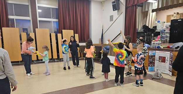Children parade indoors holding festive, colorful decorations. Some wear bright outfits, while adults guide them, creating a joyful, lively atmosphere.
