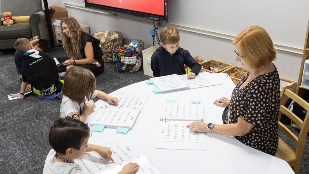 teacher sitting on floor working wih two students and teacher sitting at table working with three students