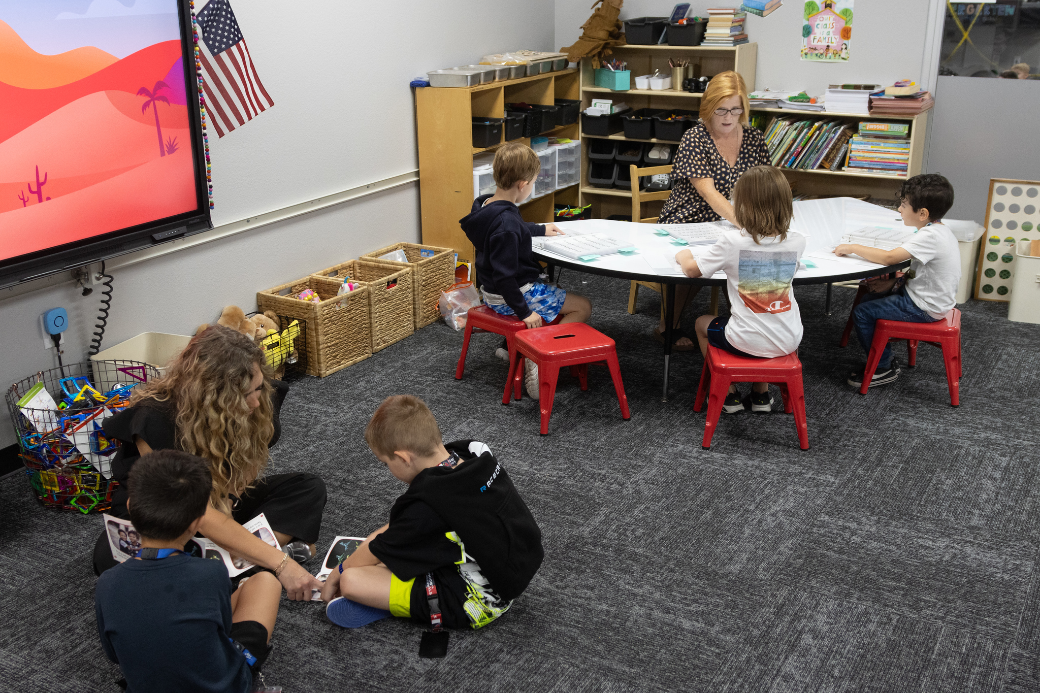 3 students work with teadher around a table, 2 students working with teacher on the floor