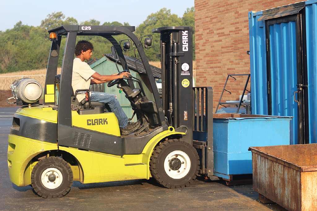 A student operates a yellow forklift to lift up a blue trash bin.