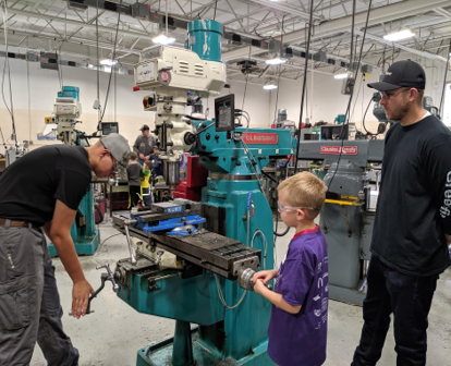 A teenage boy uses a piece of equipment in a machining lab. A young boy watches, waiting for his turn to try it out.