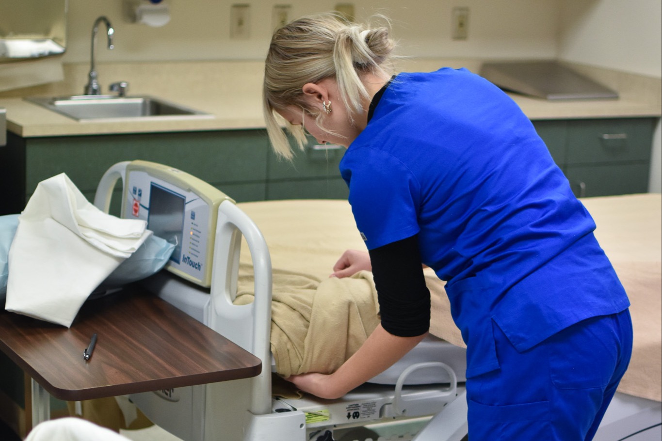 A person in blue scrubs bends over a hospital bed with a pillow and electronic monitor.