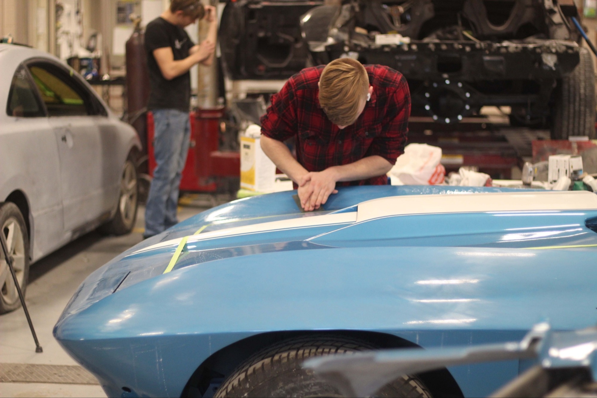 Two people in a workshop, one working on a blue car and another near a silver car.