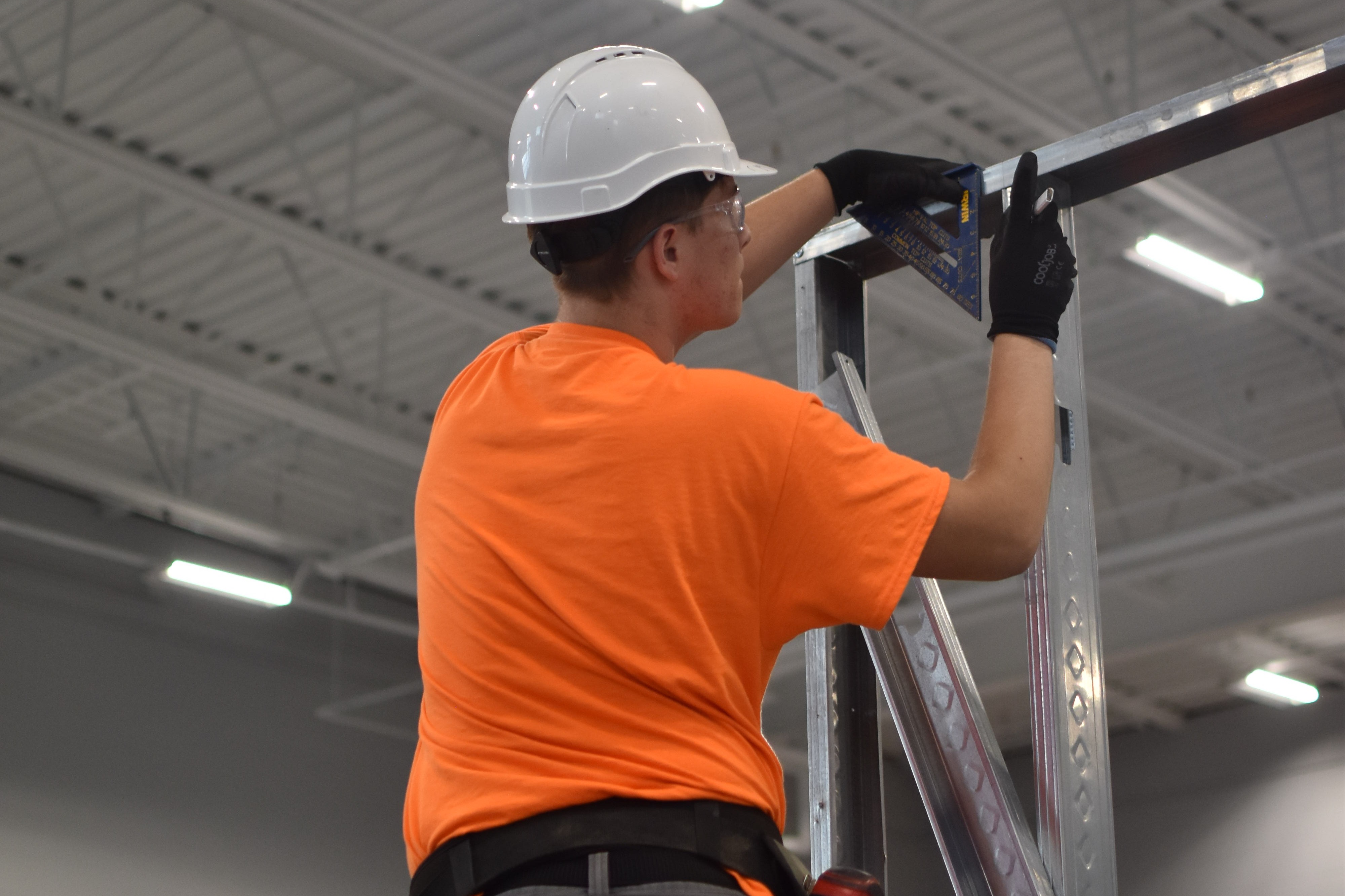 Person in an orange shirt, white hard hat, and gloves, standing on scaffolding, holding a tool.