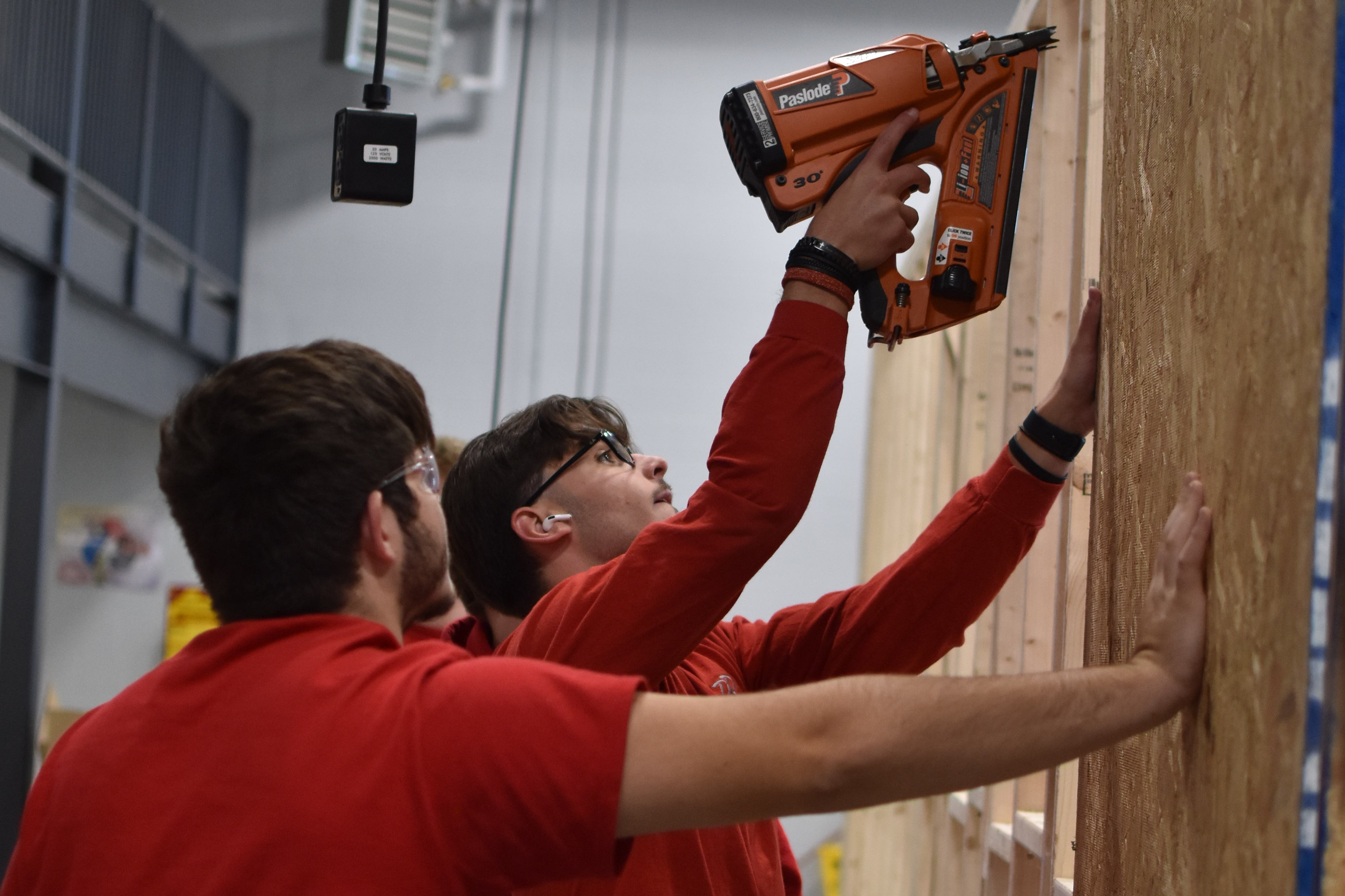 Two men in red shirts and jeans use power tools to install a wall in an unfinished room.