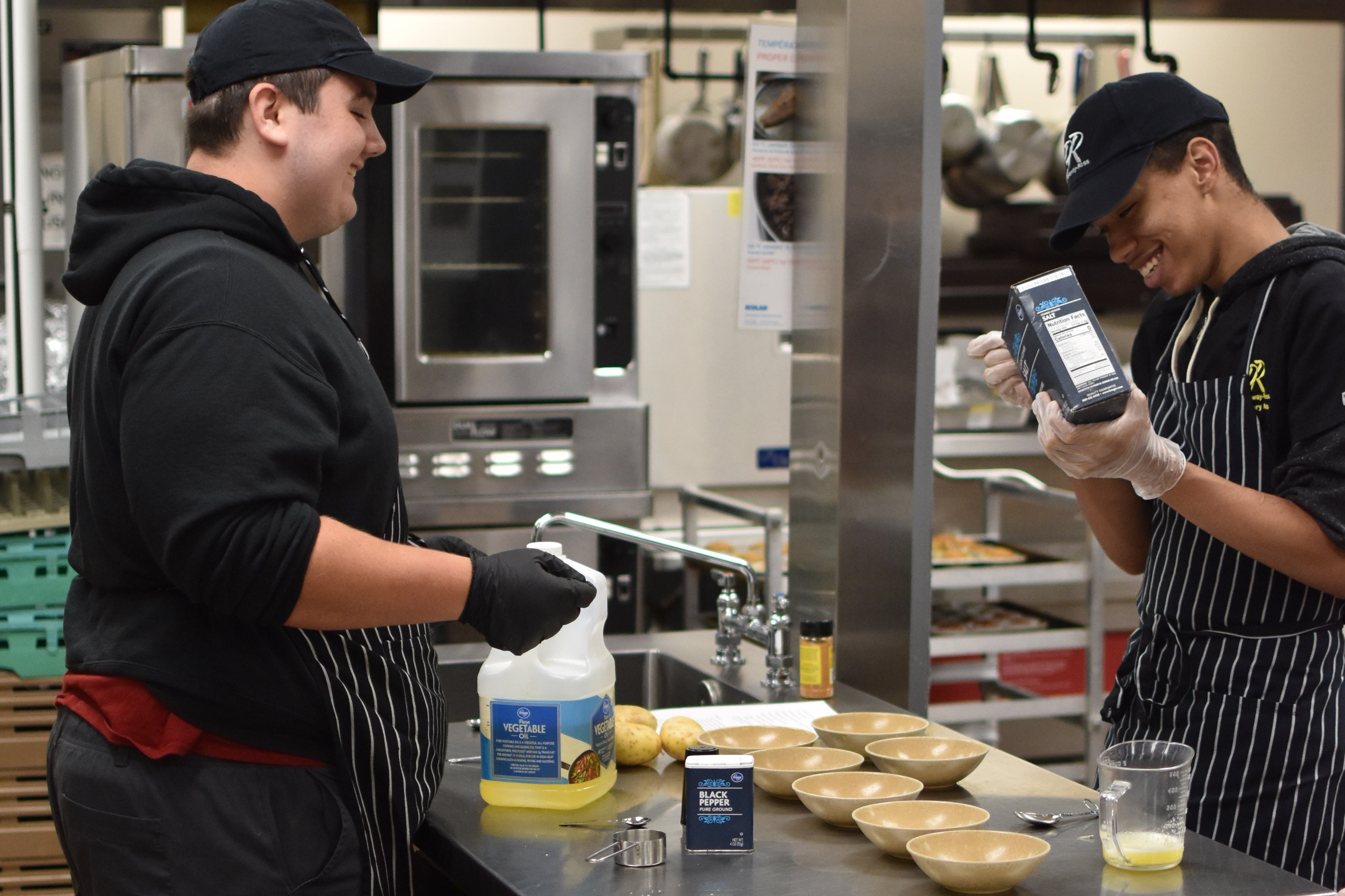 Two people in a kitchen, one holding a device, both in aprons, with bowls, plates, and utensils on the counter.