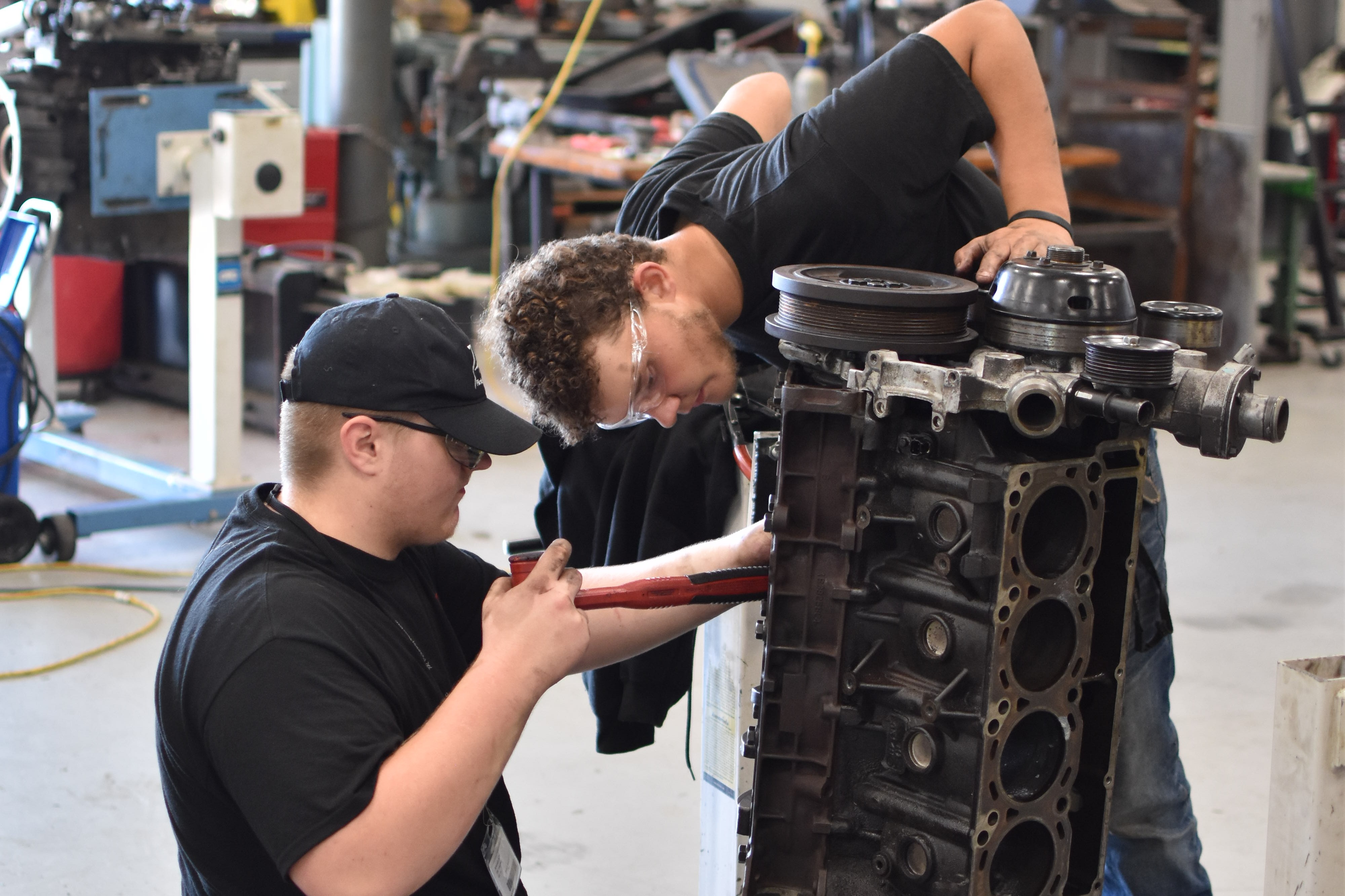 Two people wearing black t-shirts and jeans, working on an engine in a workshop with other tools and equipment.
