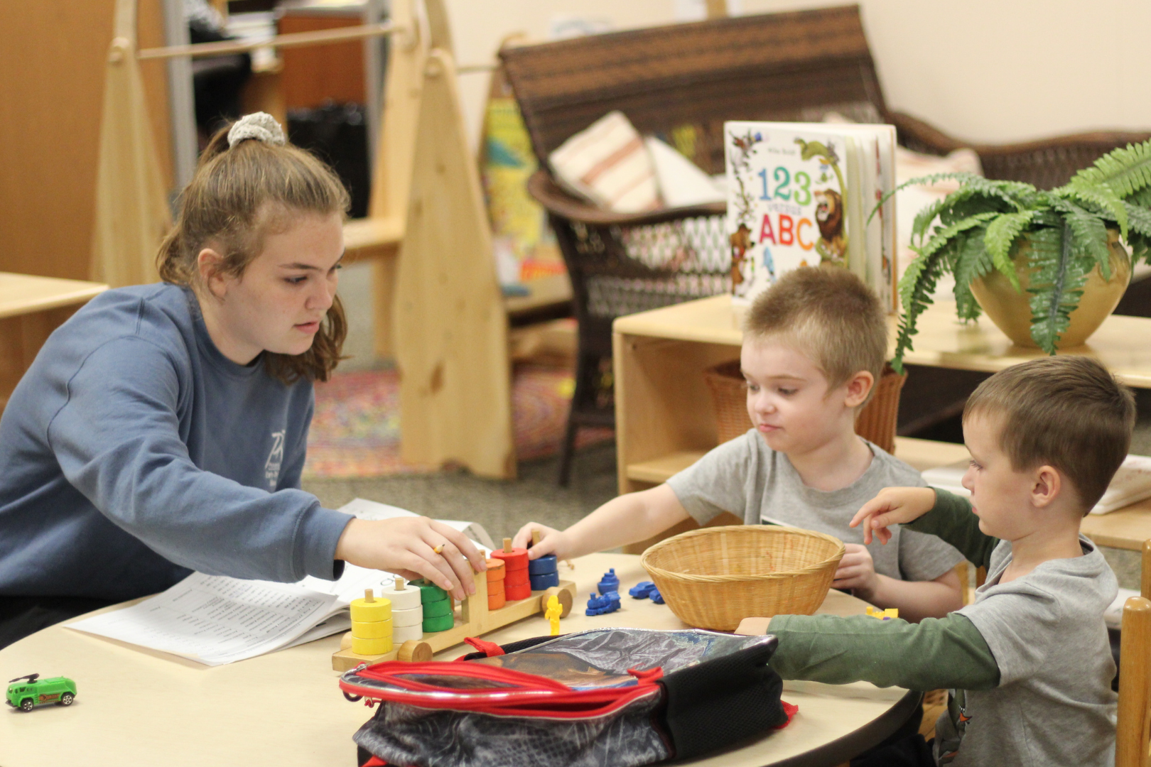 A young woman guides two young boys playing with a wooden toy at a table in a classroom.