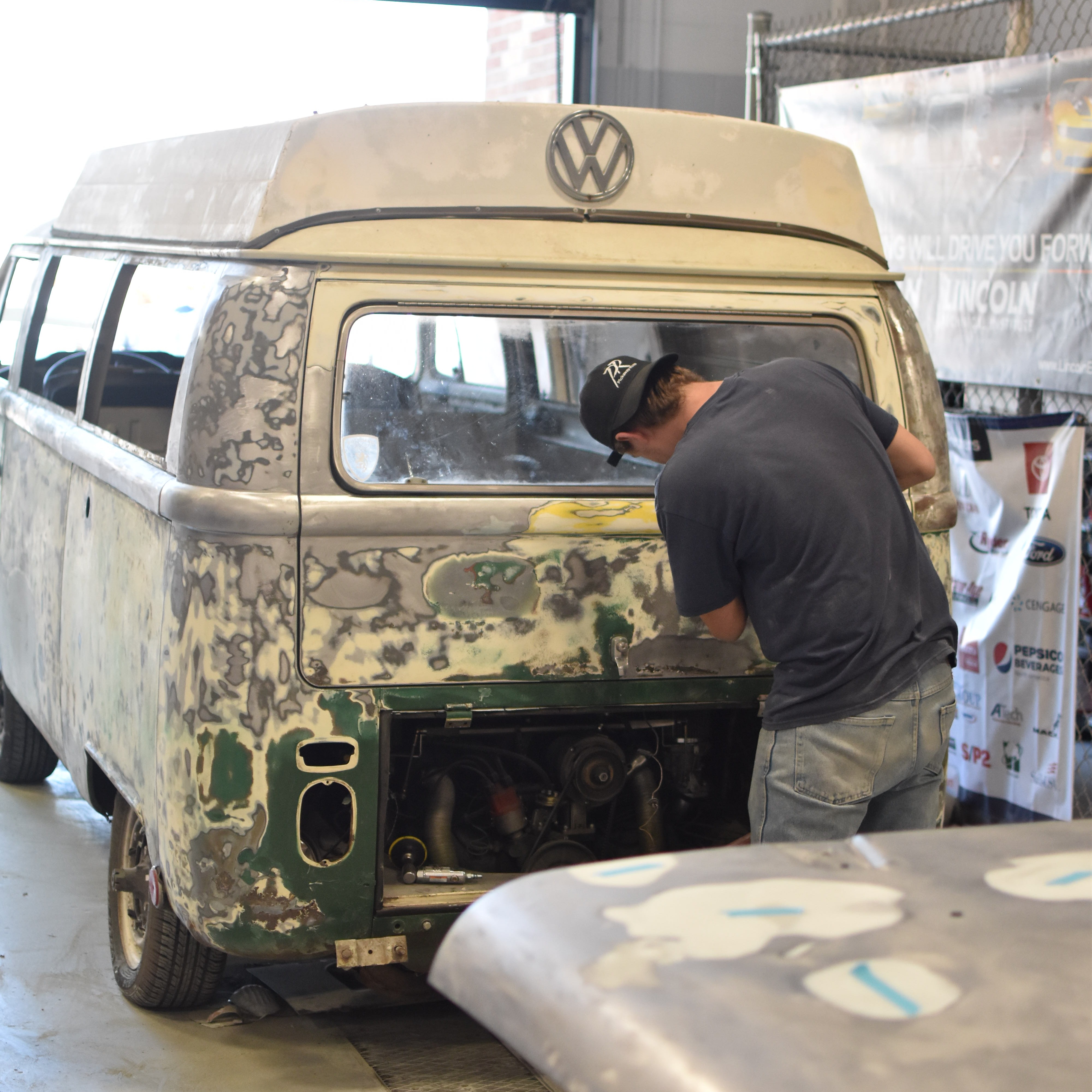 Man in a garage working on a Volkswagen van, with the back half exposed and the engine compartment open.