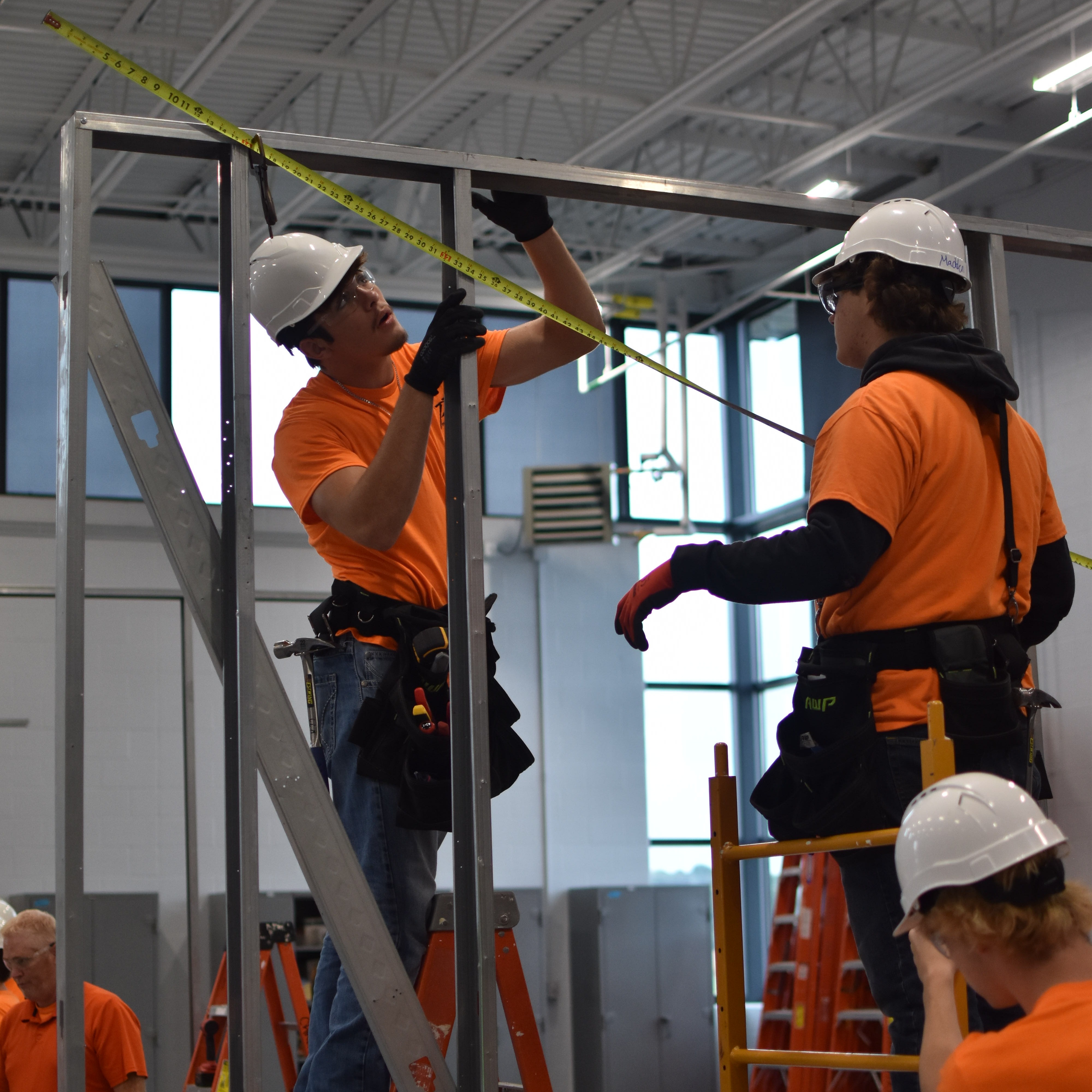 Three individuals in orange shirts and white hard hats use measuring tools in a construction area with metal frames and ladders.