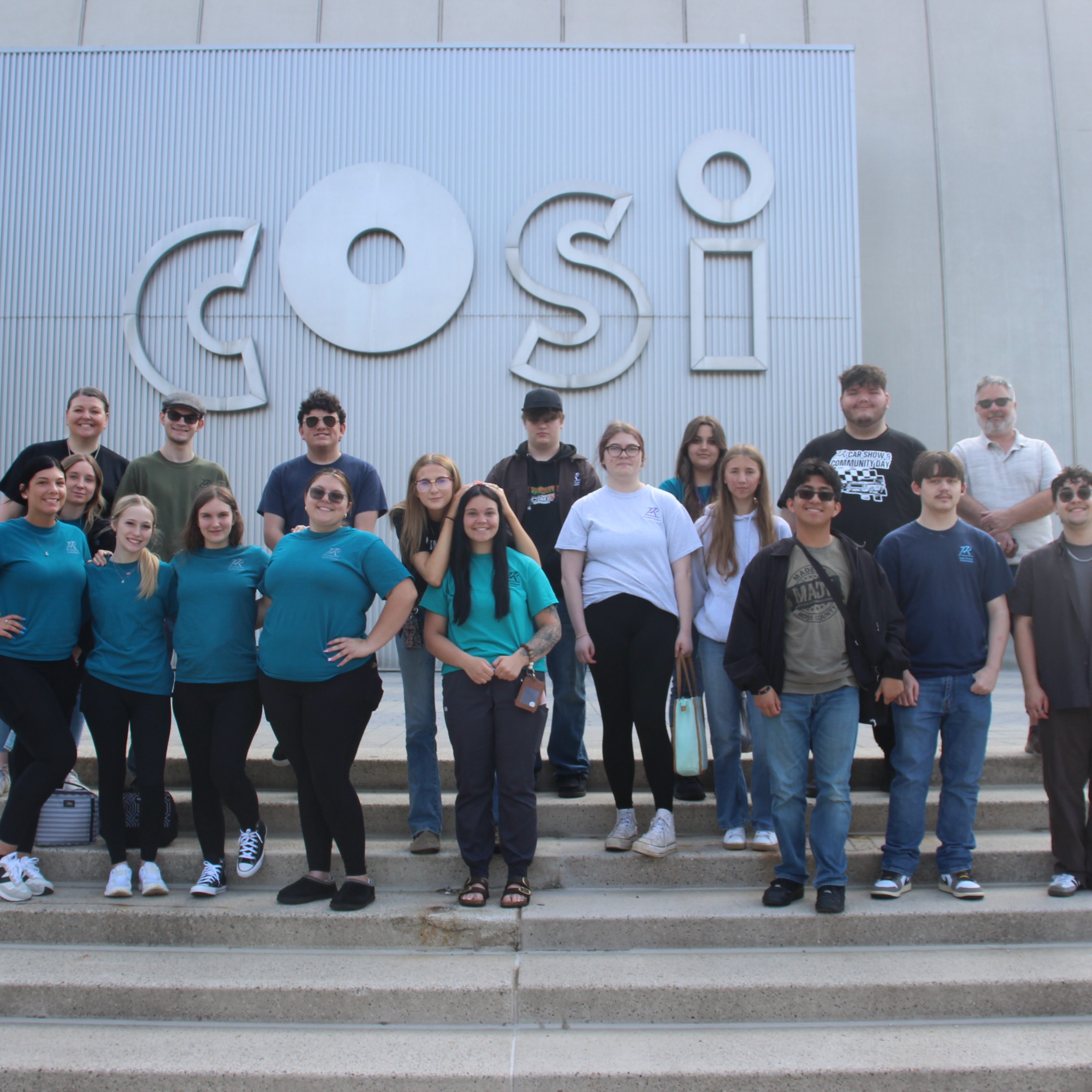 A group of people in casual clothing poses on steps in front of a white wall with a "COSI" sign.