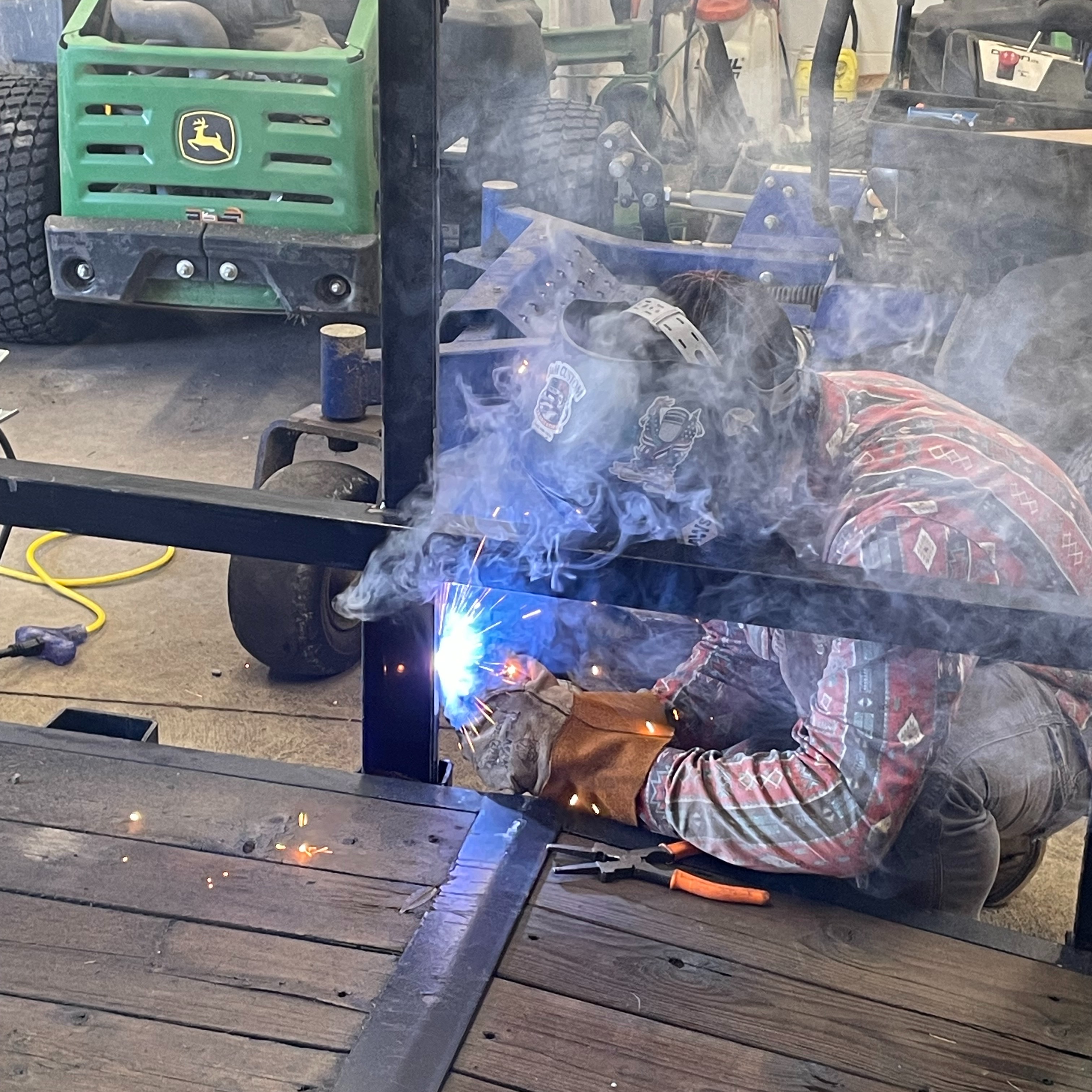 A man wearing safety gear welds metal in a garage. A green vehicle is parked in the background.