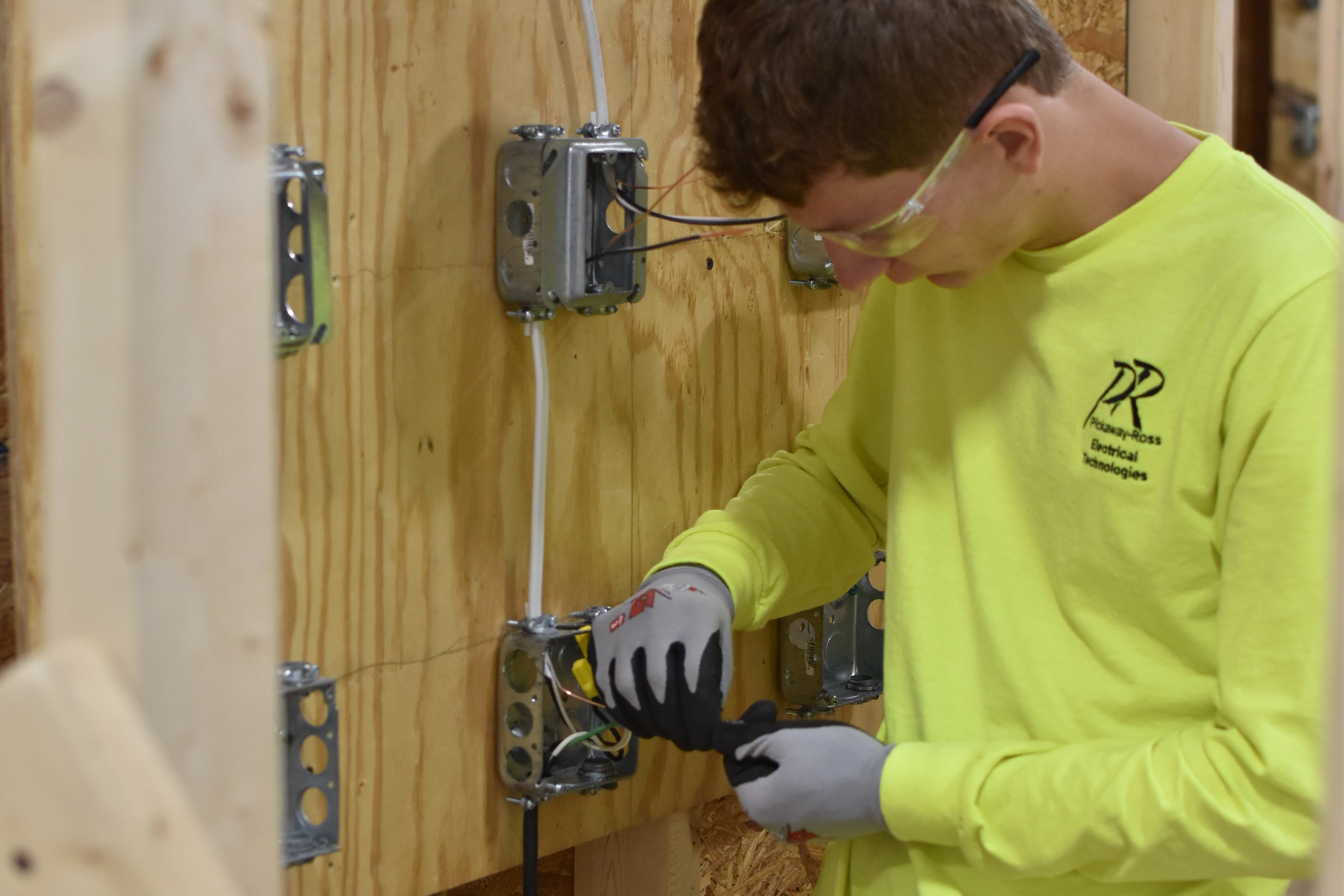 Man in a yellow shirt, glasses, and gloves works on electrical connections in a room with unfinished wooden walls.