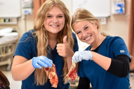 Two women in blue scrubs, gloves, and smiling, standing at a table with a mannequin torso.