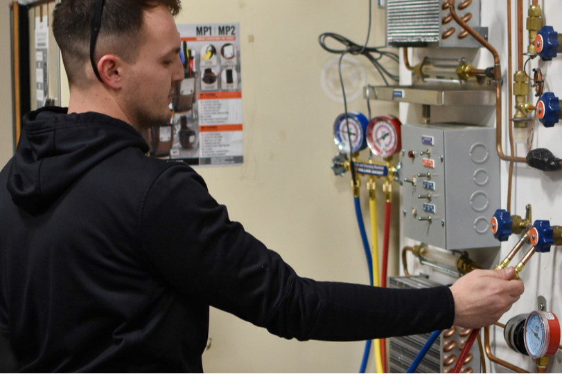 A man in a black hoodie works on a white wall with various tools and gauges.