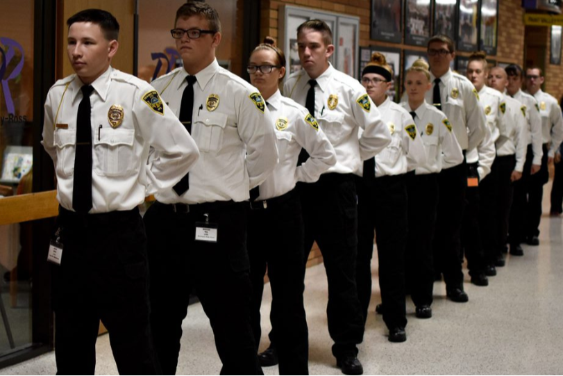 A line of security officers in white uniforms and ties walk past a glass door with a plant.