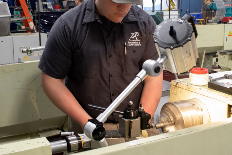 A man in a shop uses a tool to operate a machine. Behind him are other machines and metal objects.