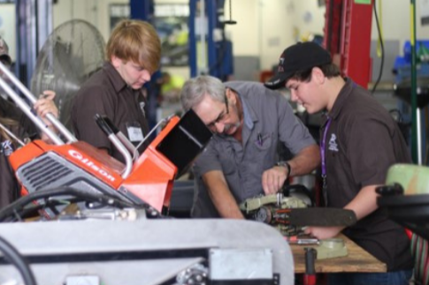 Four individuals, two male and two female, are gathered around a red machine in a garage. They are wearing uniforms.