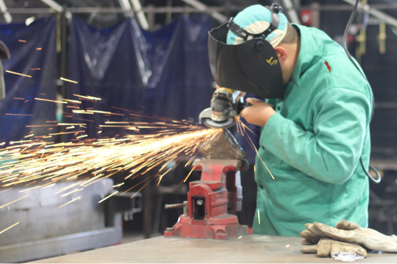 Worker wearing protective gear using an angle grinder, sparks flying. Metal tools and equipment in background.