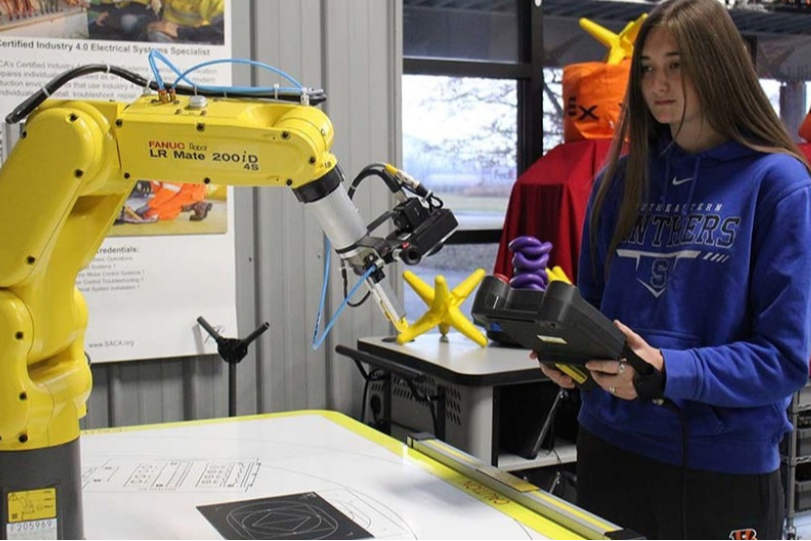 Woman in blue sweatshirt using a tablet, standing near a yellow robot. Background shows a red and white garment.