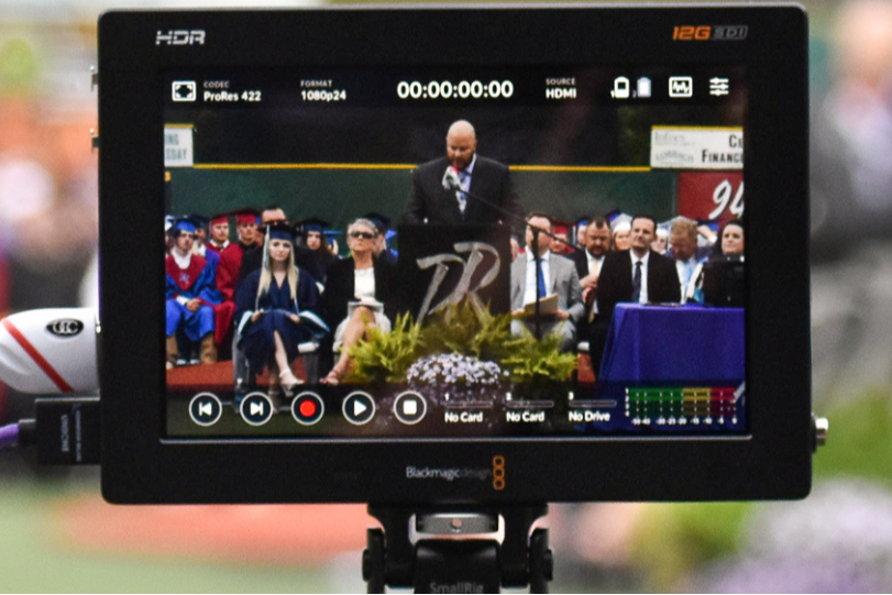 Screen of a video camera showing a man speaking to a seated audience. A microphone and cable are connected.