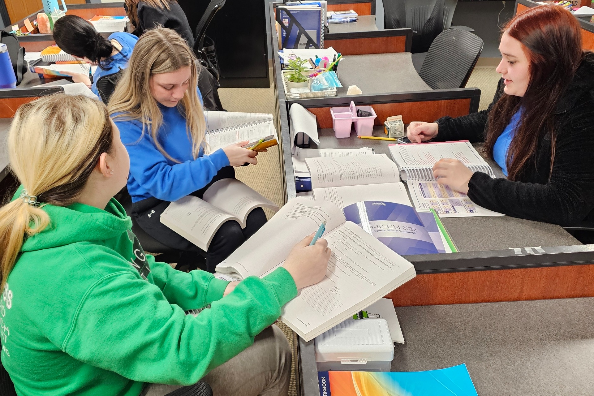 People seated in chairs around a desk, working on papers. One woman wearing a green hoodie and another wearing a blue shirt.