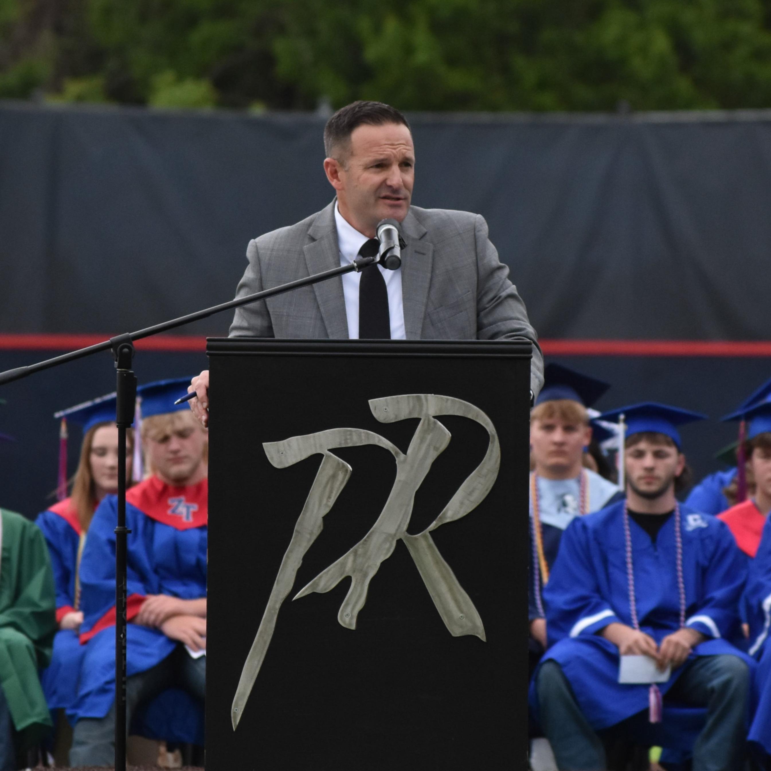 A man in a suit and tie speaks at a podium. Audience members in caps and gowns sit behind him.