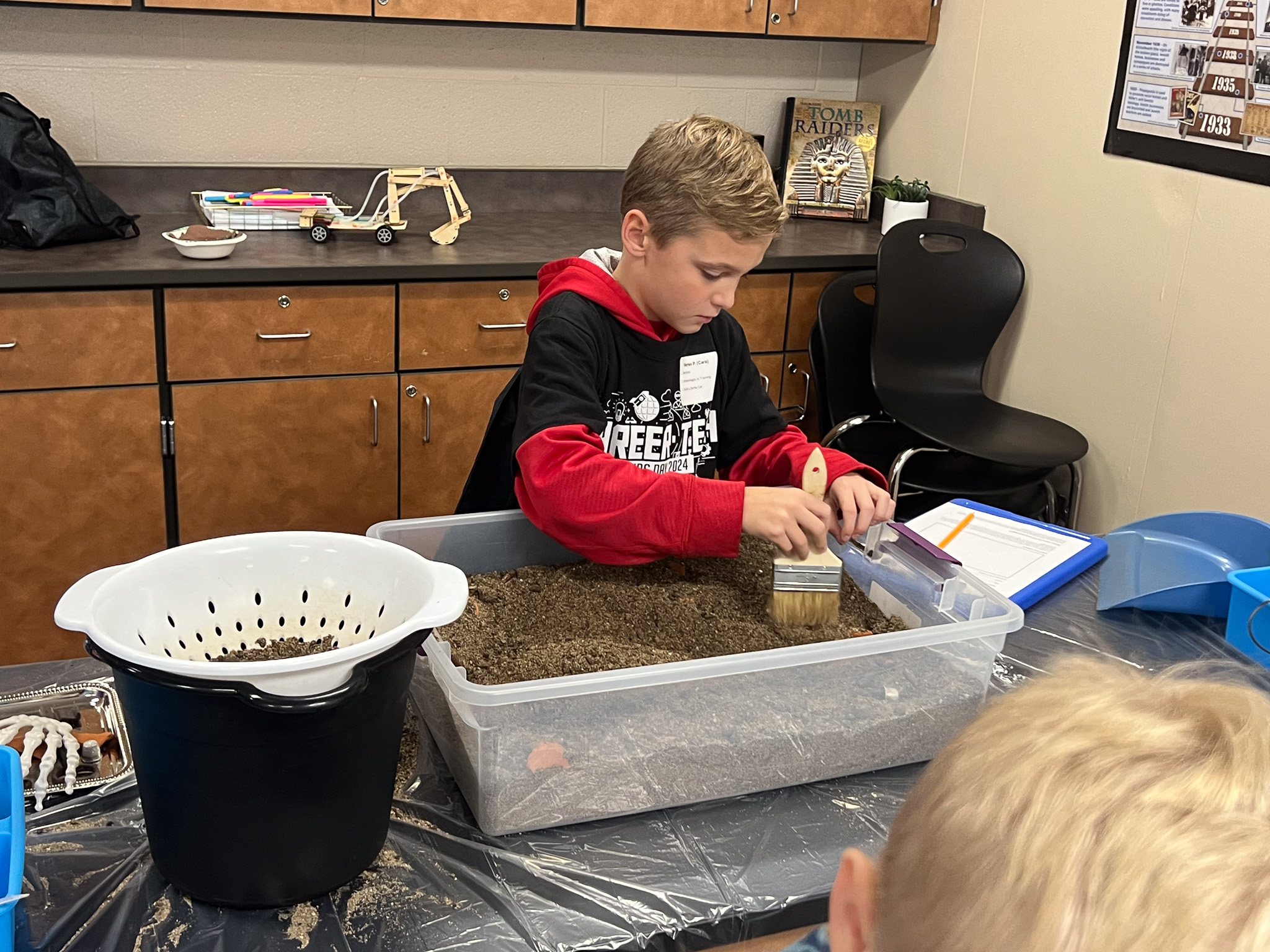 Two kids in a classroom; one uses a brush on sand in a plastic container while the other watches.
